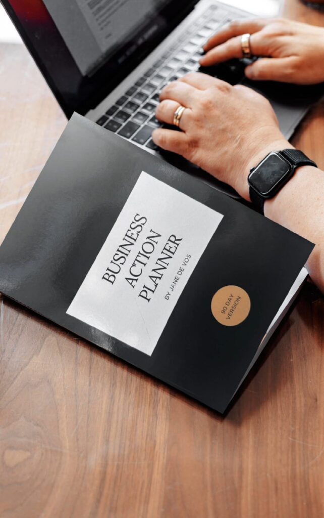 Business action planner notebook with a pen and a laptop on a white desk.