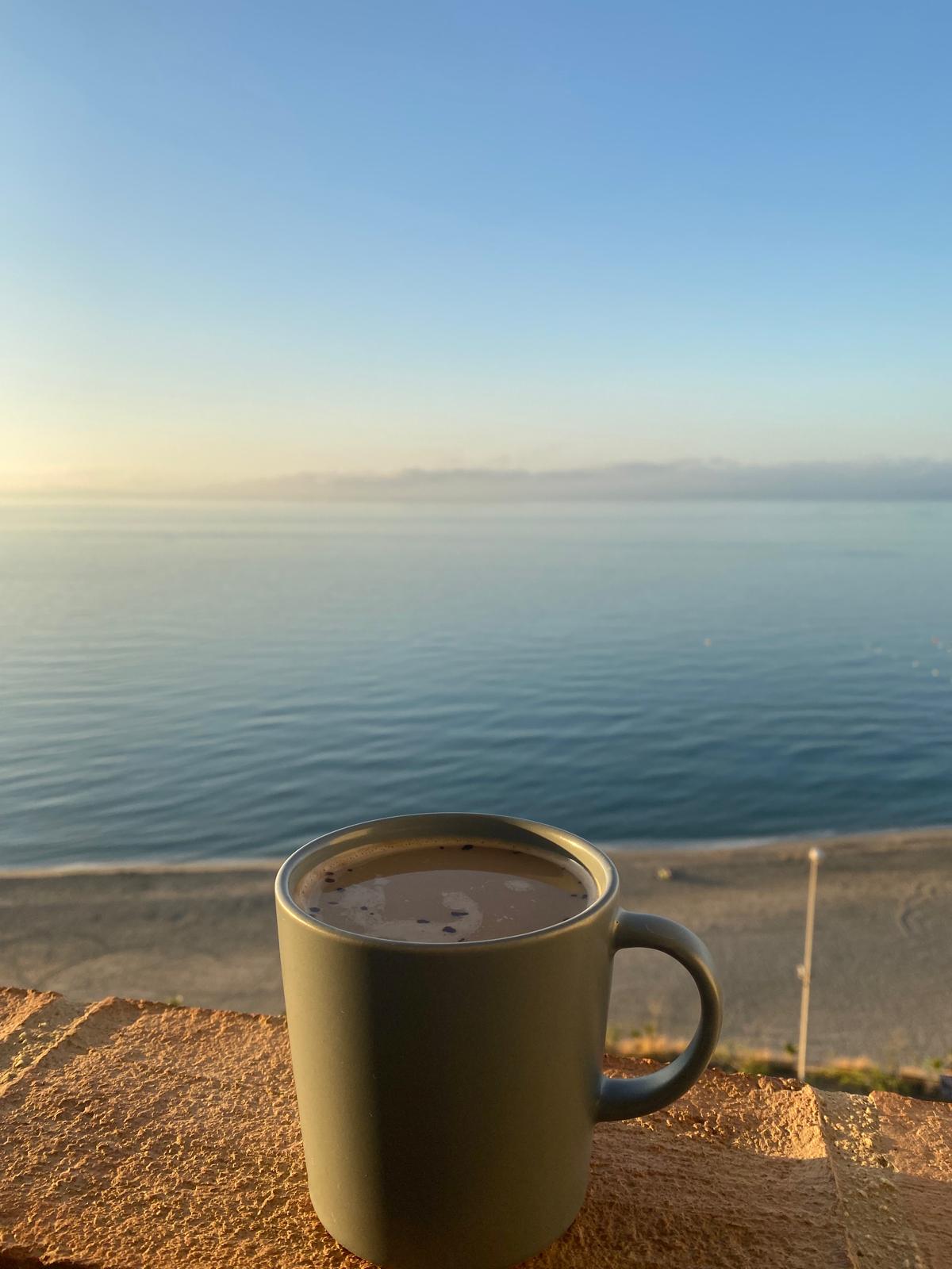 A cup of coffee placed on a wooden surface with a scenic view of the sea and sky at sunset.